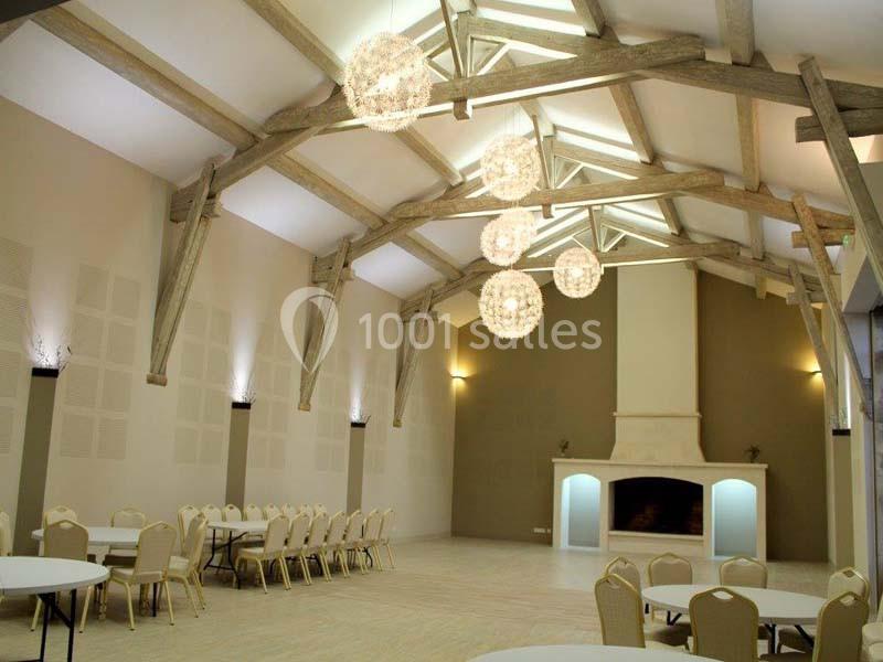 Grande salle lumineuse avec poutres apparentes, cheminée centrale, tables rondes et chaises beige clair.