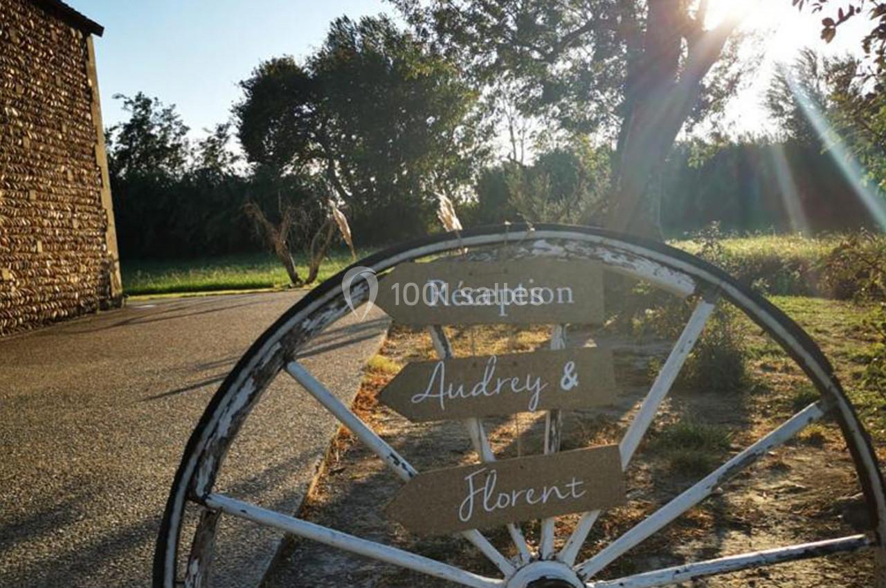 Panneau en bois indiquant ’Réception Audrey & Florent’ fixé sur une roue, dans un cadre champêtre ensoleillé.