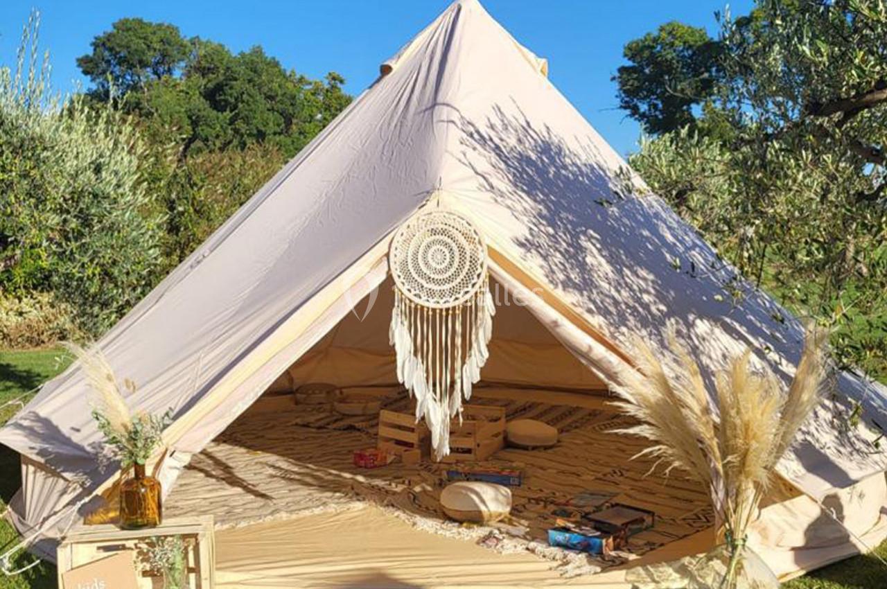 Tente beige en toile installée sur une pelouse, décorée avec un macramé, des coussins et des plantes sèches.