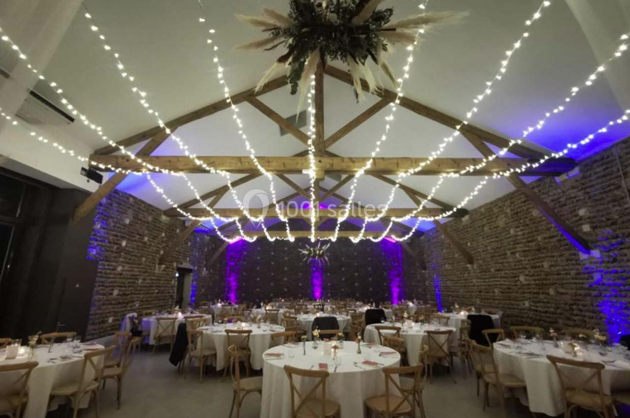 Salle de réception avec murs en pierre, tables dressées et guirlandes lumineuses suspendues au plafond en bois.