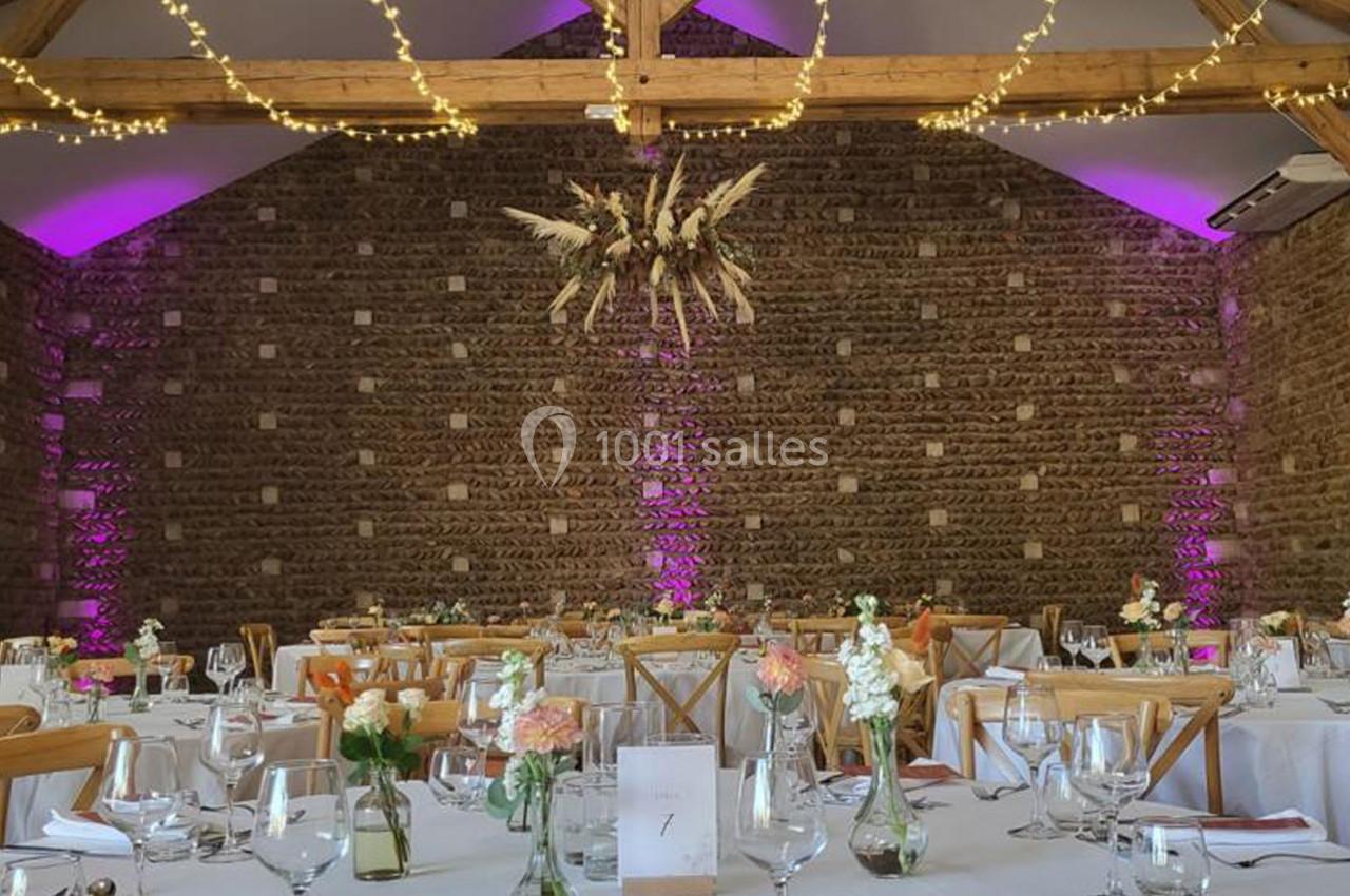 Salle de réception en pierre avec tables décorées, chaises en bois, guirlandes lumineuses et éclairage violet.
