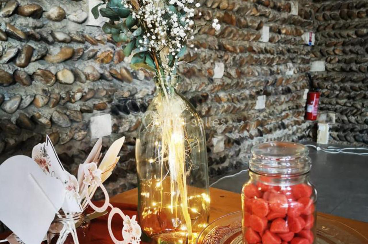 Vase en verre avec guirlande lumineuse, fleurs séchées, pot de bonbons rouges et décor en papier sur une table en bois.