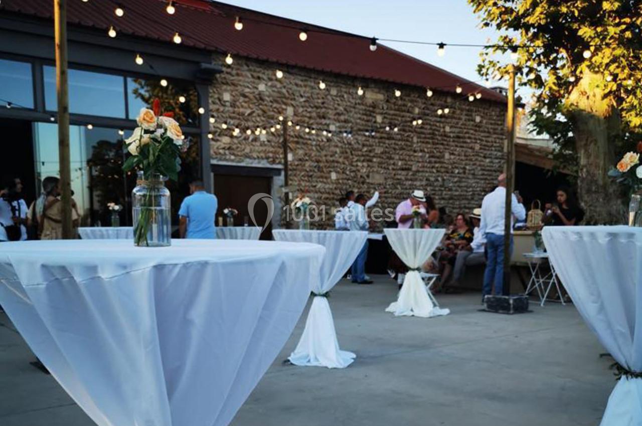 Tables hautes décorées de nappes blanches et de fleurs, disposées en extérieur sous des guirlandes lumineuses.