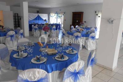Salle de réception décorée avec des tables dressées, nappes blanches, chaises ornées de nœuds violets et centres de table…