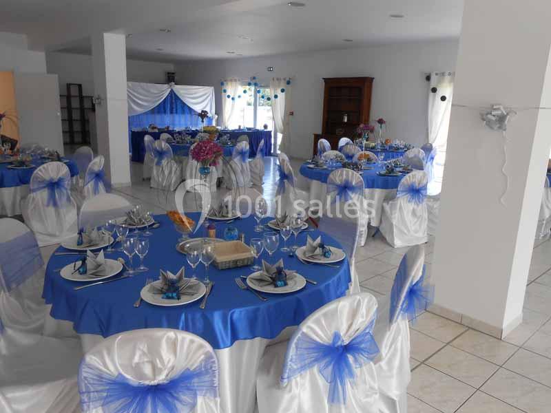 Salle décorée pour un événement avec des tables rondes nappées de bleu, chaises blanches ornées de nœuds bleus.