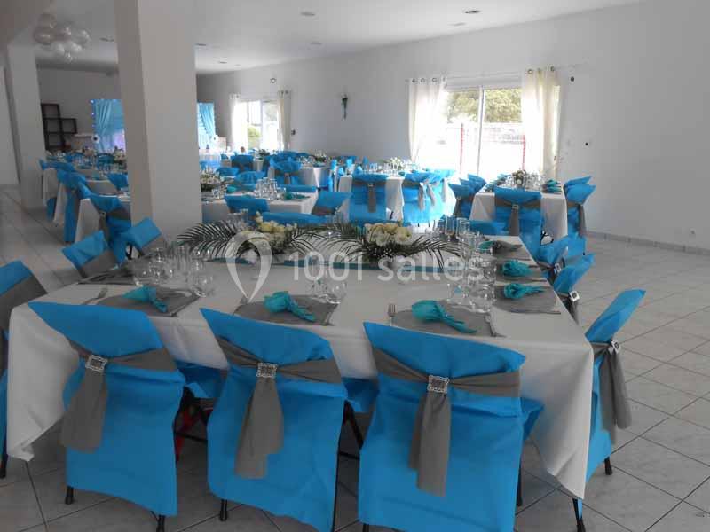 Salle de réception décorée avec des tables dressées, nappes blanches et chaises couvertes de housses bleues et grises.