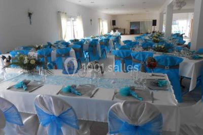 Salle de réception décorée avec des tables dressées, nappes blanches, chaises ornées de nœuds violets et centres de table…