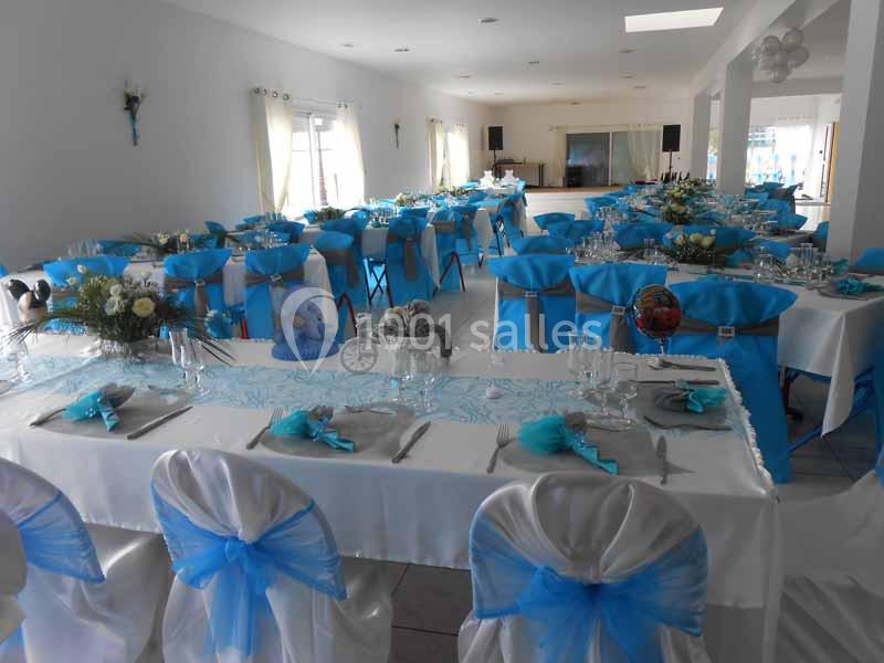 Salle de réception décorée avec des nappes blanches et des chaises ornées de nœuds bleus, tables dressées pour un événement.