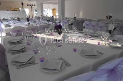 Salle de réception décorée avec des tables dressées, nappes blanches, chaises ornées de nœuds violets et centres de table…
