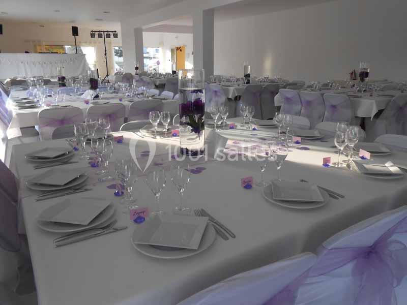 Salle de réception décorée avec des tables dressées, nappes blanches, chaises ornées de nœuds violets et centres de table…