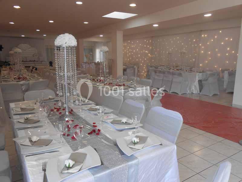 Salle de réception décorée avec des tables dressées, nappes blanches, chemins de table argentés et guirlandes lumineuses.