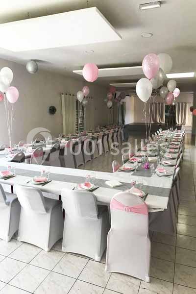Salle décorée pour une réception, avec des tables alignées, nappes blanches, chaises couvertes et ballons roses et blancs.