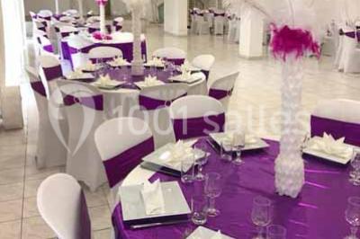 Salle de réception décorée avec des tables dressées, nappes blanches, chaises ornées de nœuds violets et centres de table…