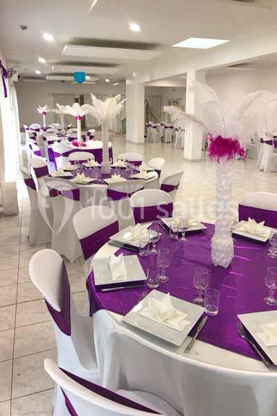 Salle de réception décorée avec des tables rondes, nappes violettes, chaises blanches et centres de table ornés de plumes…