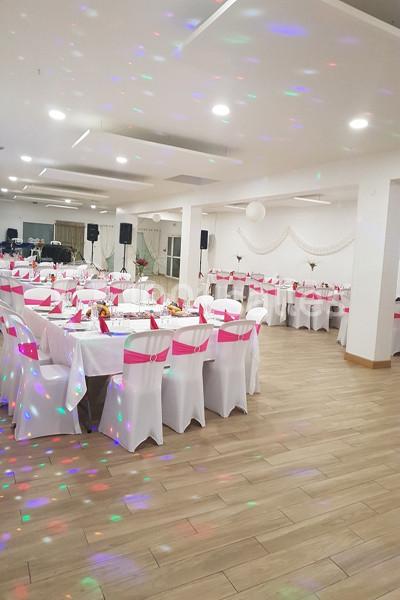 Salle de réception décorée avec des nappes blanches et des rubans roses, éclairée par des lumières colorées.