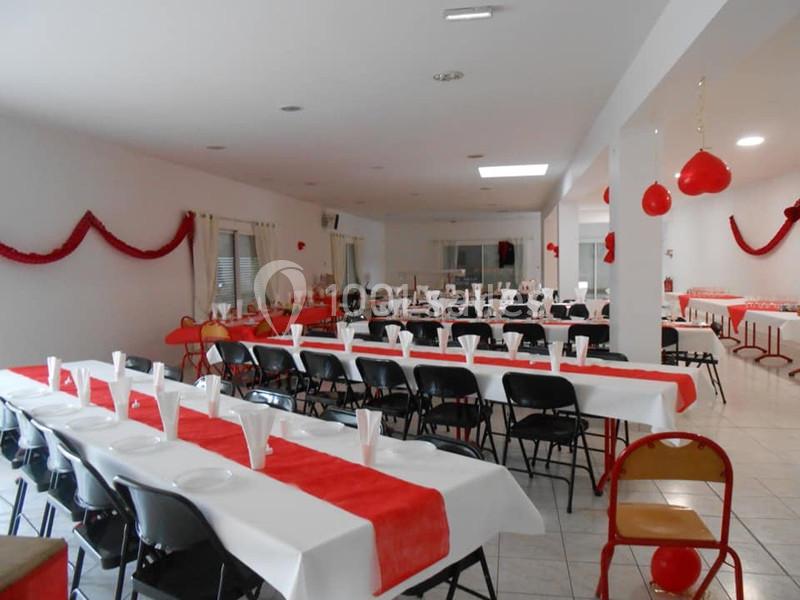 Salle décorée avec des nappes blanches et rouges, tables alignées avec chaises noires, prête pour un événement.
