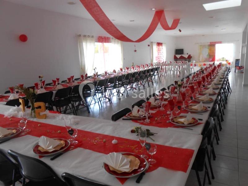Salle décorée pour un événement, avec de longues tables alignées, nappes blanches et rouges, et vaisselle dressée.