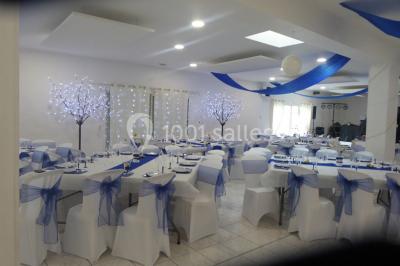 Salle de réception décorée avec des tables dressées, nappes blanches, chaises ornées de nœuds violets et centres de table…