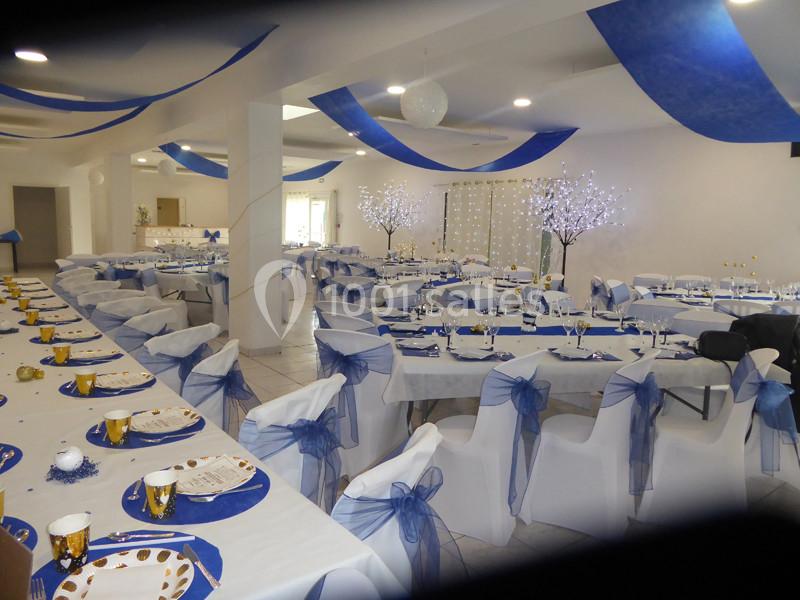 Salle de réception décorée en blanc et bleu avec des tables dressées, des chaises ornées de nœuds et des arbres lumineux.