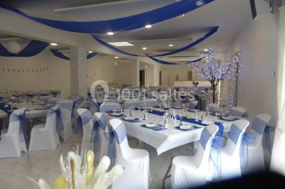 Salle de réception décorée avec des tables dressées, nappes blanches, chaises ornées de nœuds violets et centres de table…