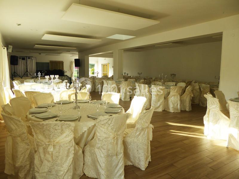 Salle de réception lumineuse avec des tables rondes dressées, nappes et chaises recouvertes de tissu beige décoratif.
