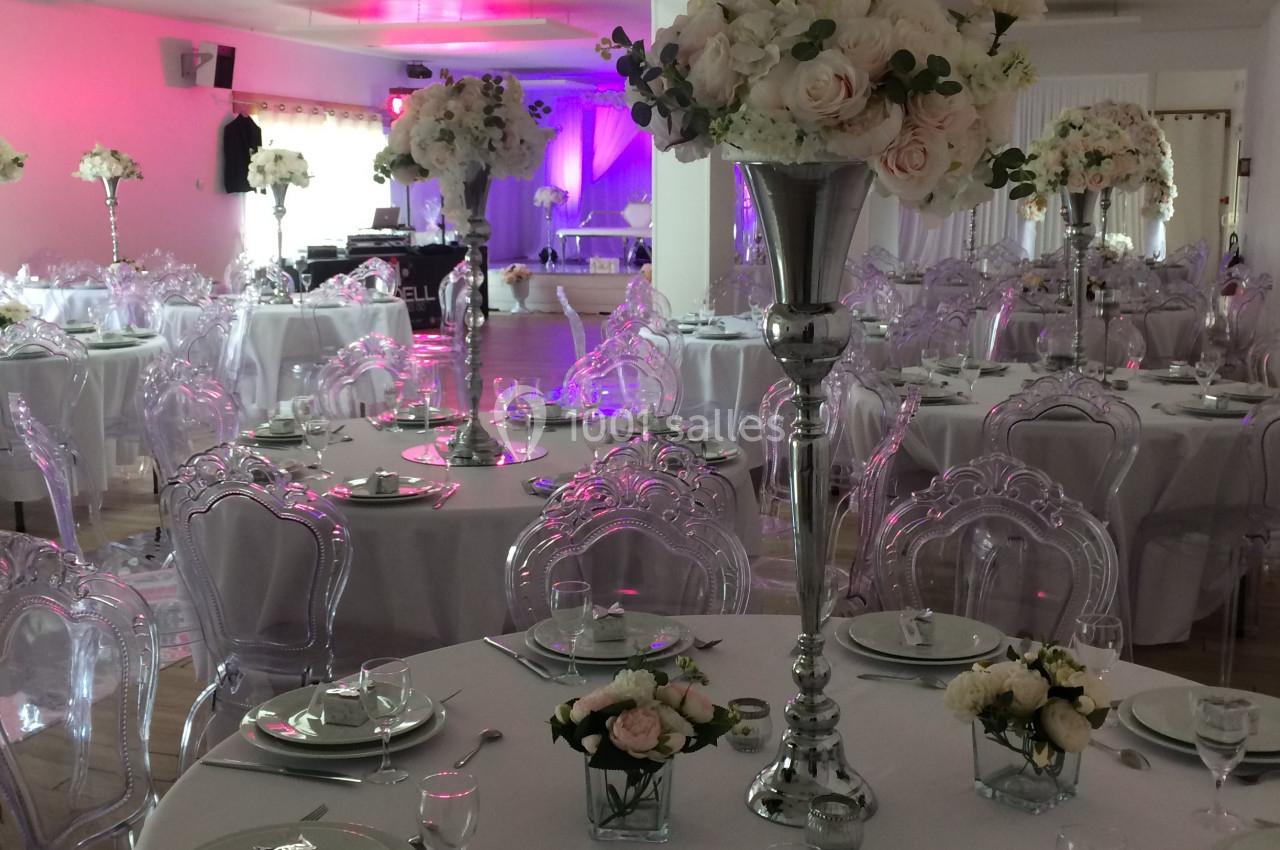 Salle de réception décorée avec des tables rondes, nappes blanches, centres de table floraux et chaises transparentes.