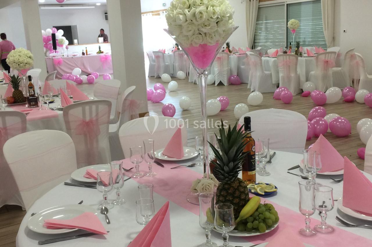 Salle décorée pour un événement avec tables dressées, nappes blanches, serviettes roses et ballons roses et blancs.