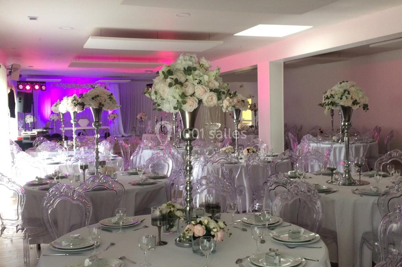 Salle de réception décorée avec des tables rondes, nappes blanches, chaises transparentes et centres de table fleuris.