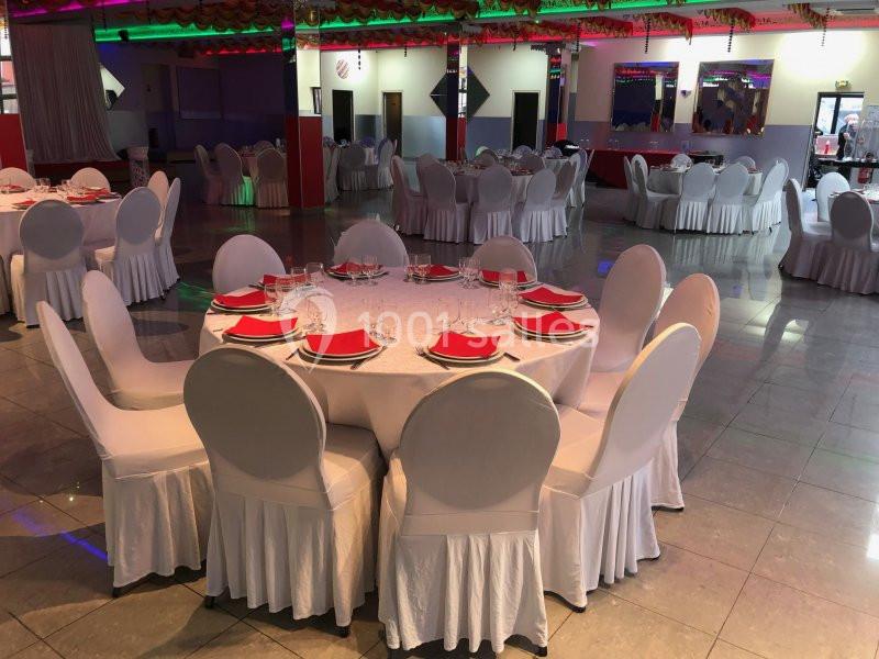 Salle de réception avec tables rondes dressées, nappes blanches, serviettes rouges et chaises couvertes de housses blanches.