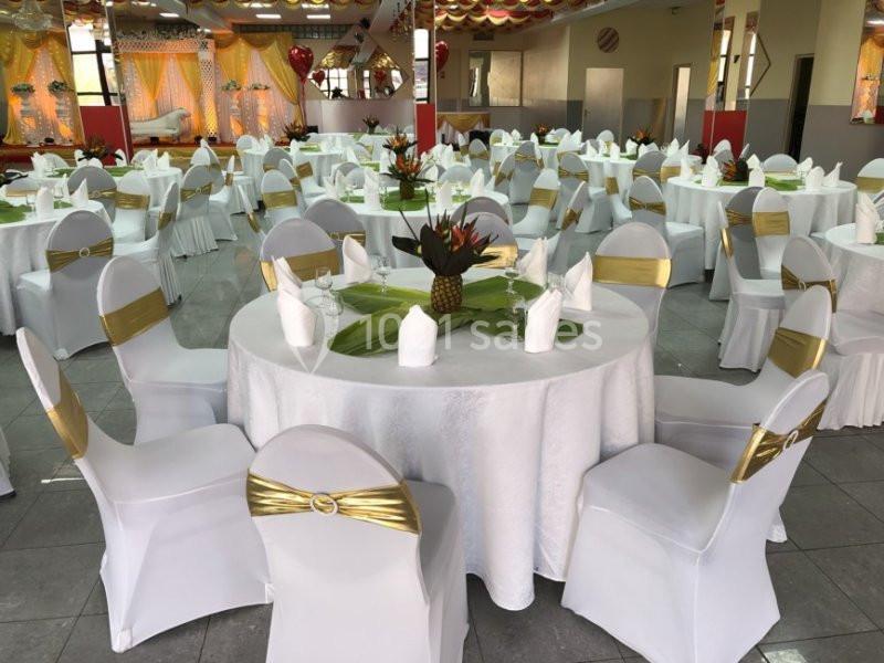 Salle décorée pour un événement avec des tables rondes dressées, nappes blanches et chaises ornées de rubans dorés.