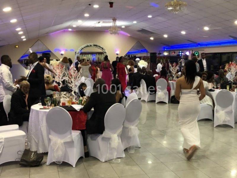 Salle de réception animée avec des invités en tenue élégante, assis à des tables décorées de nappes blanches.