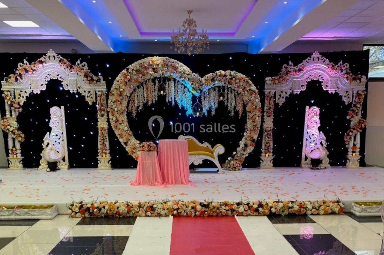 Décor de scène de mariage avec arche en forme de cœur ornée de fleurs, chaise dorée et éclairage lumineux.