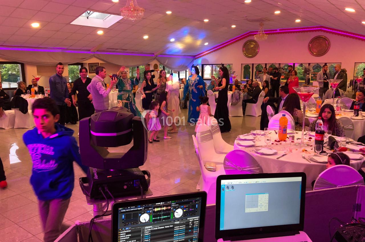 Salle de réception animée avec des invités dansant et dînant, vue depuis une table de DJ équipée d'ordinateurs.