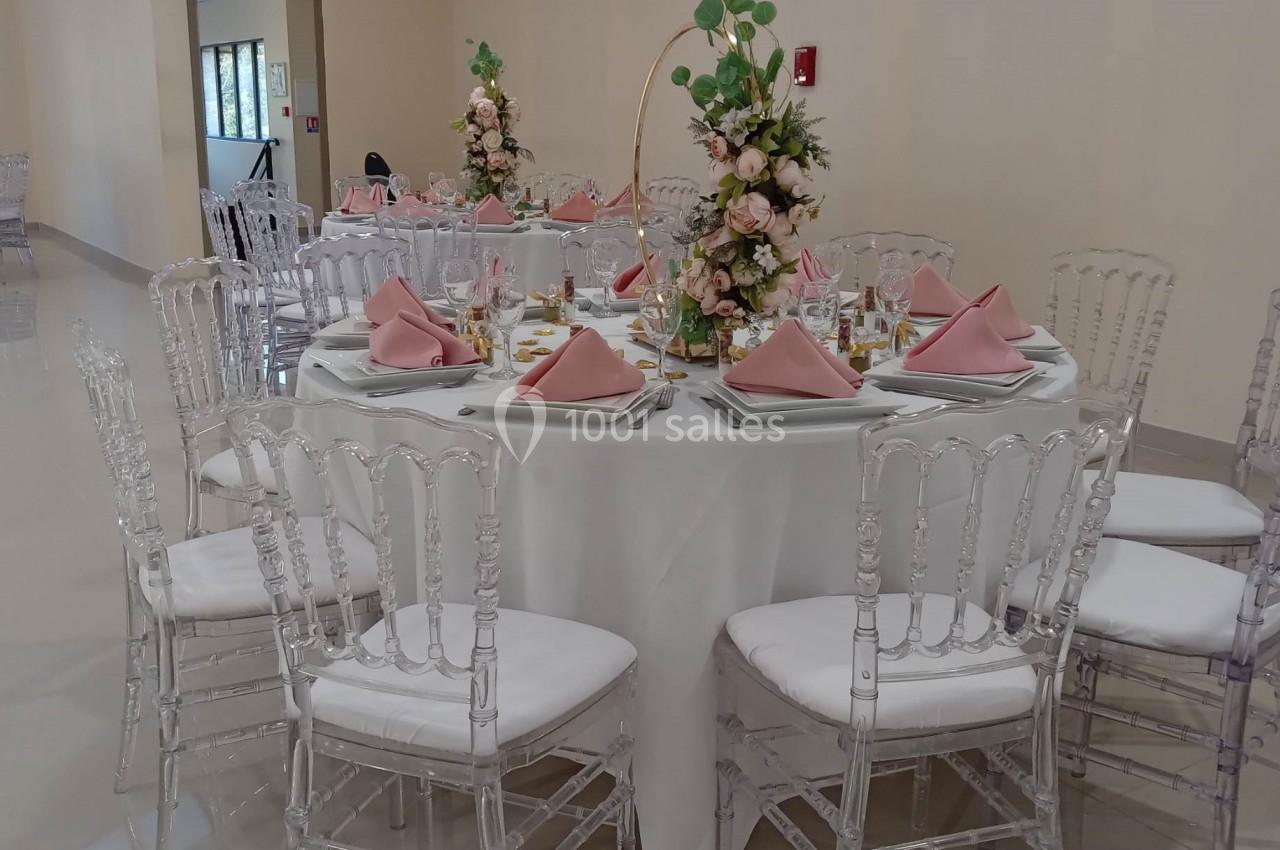 Table ronde décorée pour un événement, avec nappes blanches, serviettes roses, centres de table fleuris et chaises…