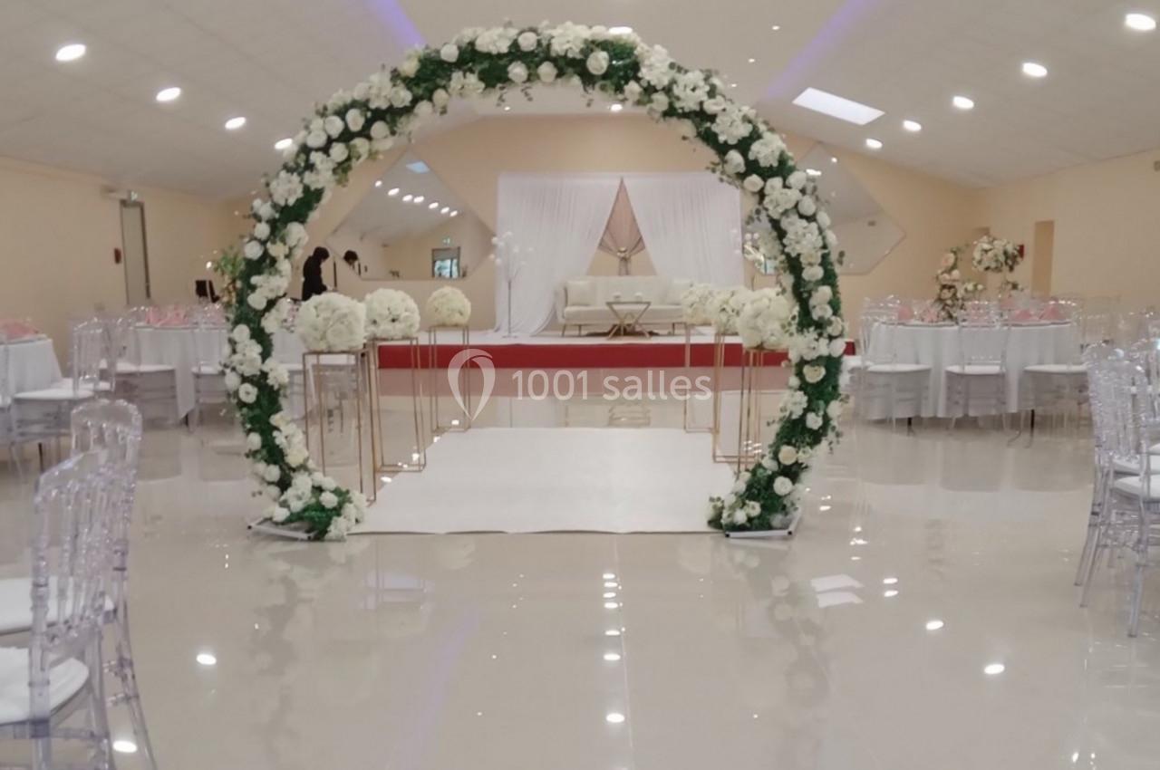 Salle de réception décorée avec des tables dressées, un arche floral et un espace central aménagé pour une cérémonie.