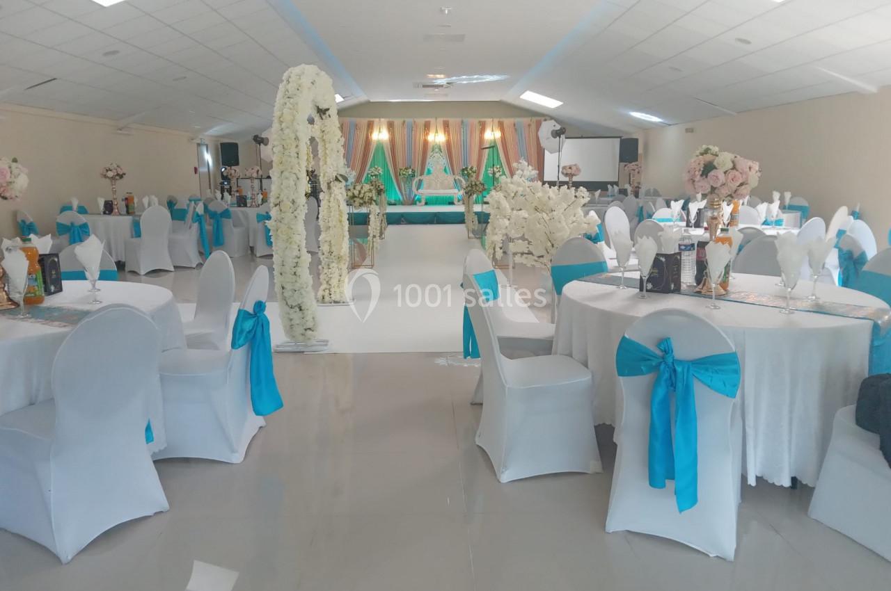 Salle de réception décorée avec des tables rondes, nappes blanches, chaises ornées de rubans bleus et arches fleuries.