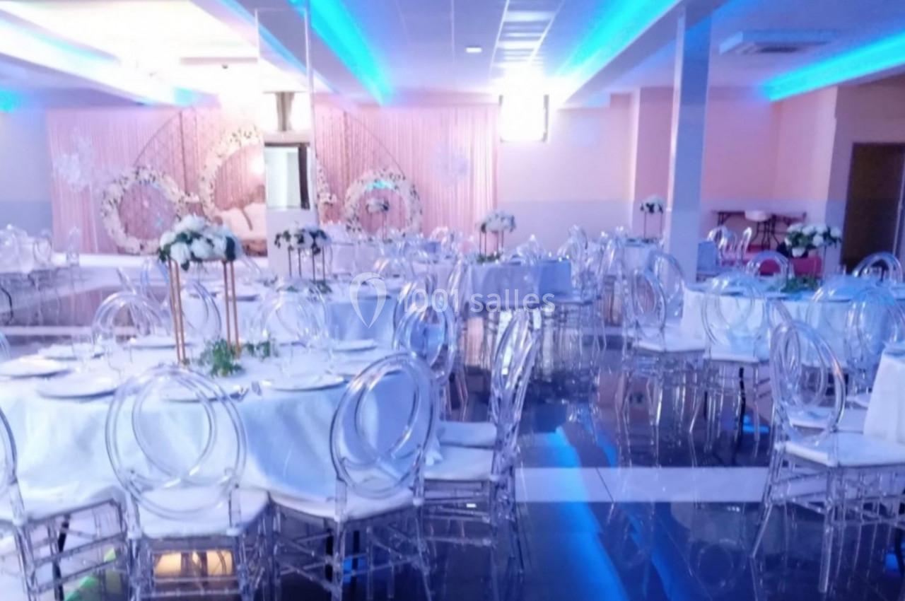 Salle de réception décorée avec des tables rondes, nappes blanches, chaises transparentes et éclairage bleu.