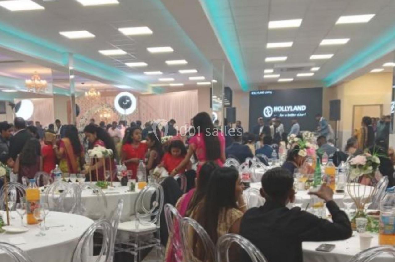 Salle de réception animée avec invités assis et debout, tables décorées de fleurs et éclairage lumineux.