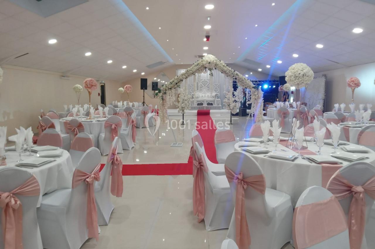 Salle de réception décorée pour un mariage avec des tables dressées, des chaises ornées de nœuds roses et une arche fleurie.