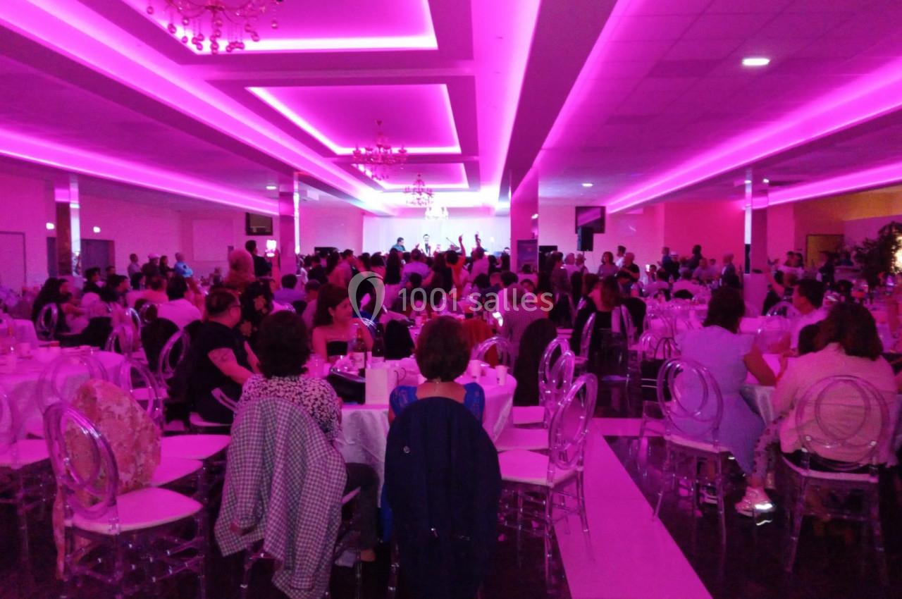 Salle de réception éclairée en rose, remplie de personnes assises autour de tables, regardant une scène au fond.