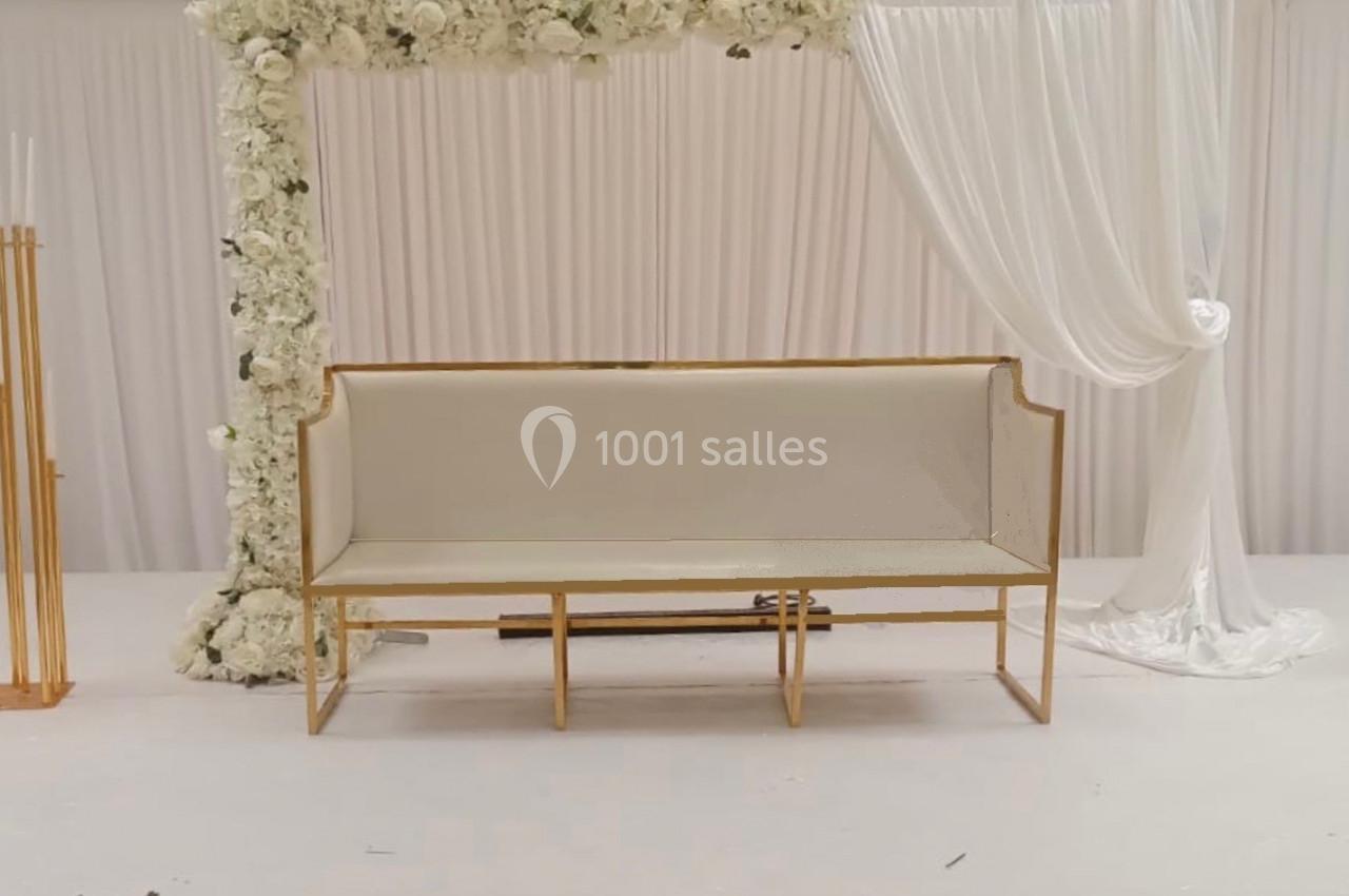 Banc beige devant une arche décorée de fleurs blanches et de rideaux, entouré de chandeliers dorés.