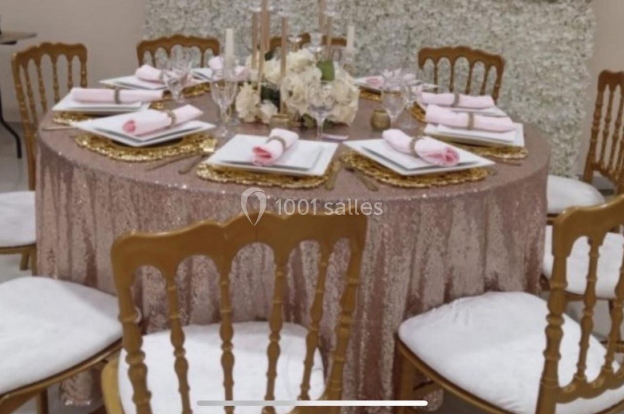 Table ronde décorée avec une nappe rose scintillante, des chaises dorées, des assiettes blanches et un centre de table…