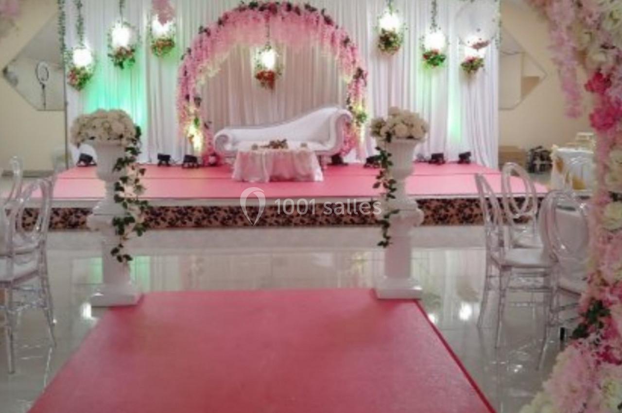 Salle de réception décorée pour un mariage avec arche florale, estrade rose, canapé blanc et arrangements floraux.