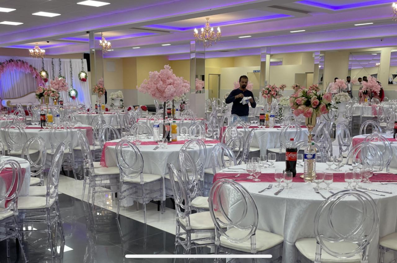 Salle de réception décorée avec des tables rondes, nappes blanches, chaises transparentes et arrangements floraux roses.