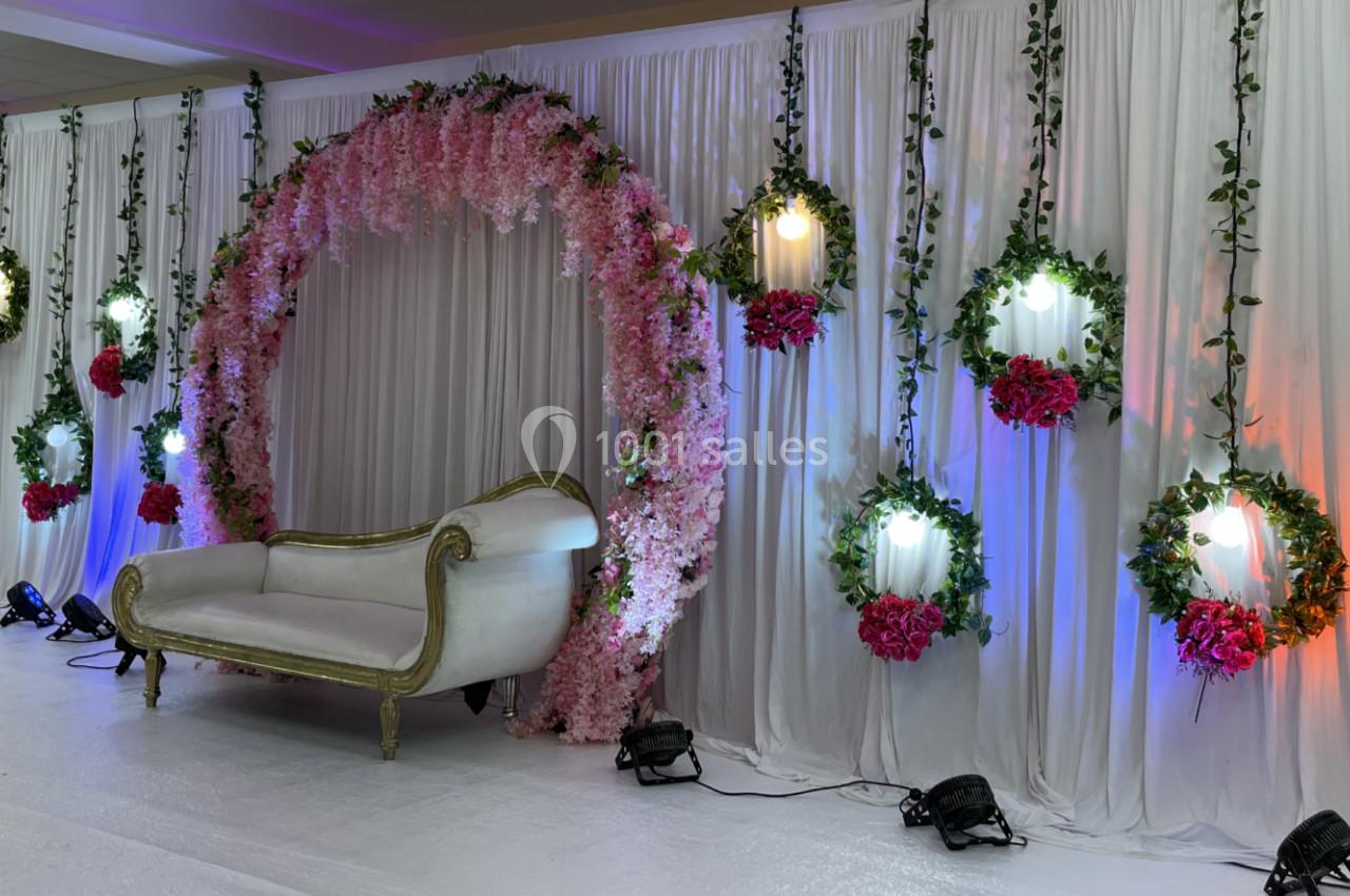 Décor de cérémonie avec arche florale rose, canapé blanc et guirlandes lumineuses entourées de fleurs.