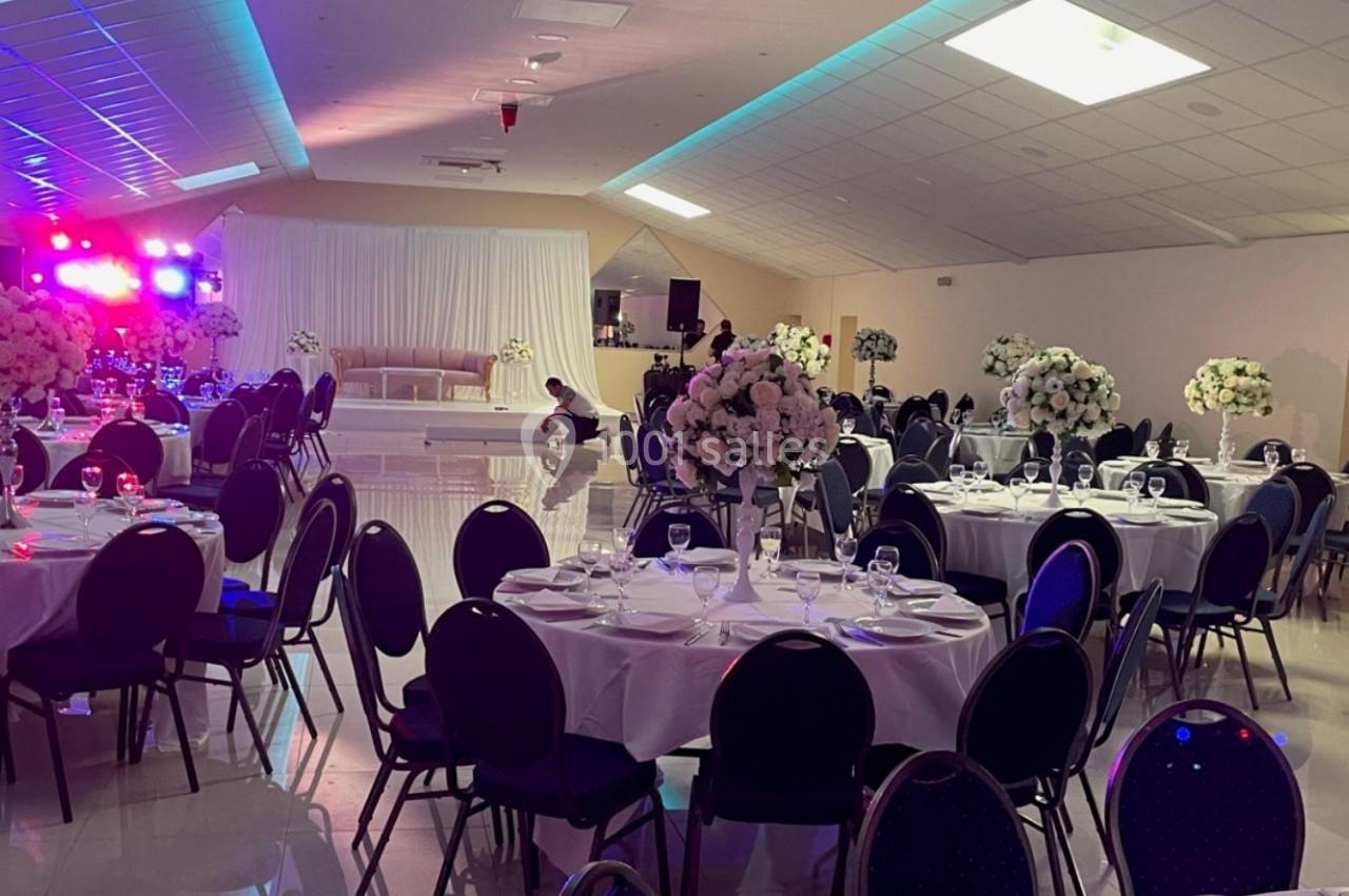 Salle de réception décorée avec des tables rondes, chaises noires et arrangements floraux, éclairage coloré en arrière-plan.