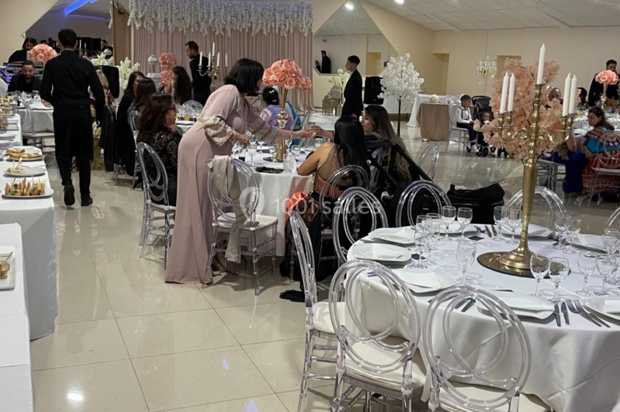 Salle de réception décorée avec des tables rondes, des chandeliers et des invités assis ou se servant au buffet.
