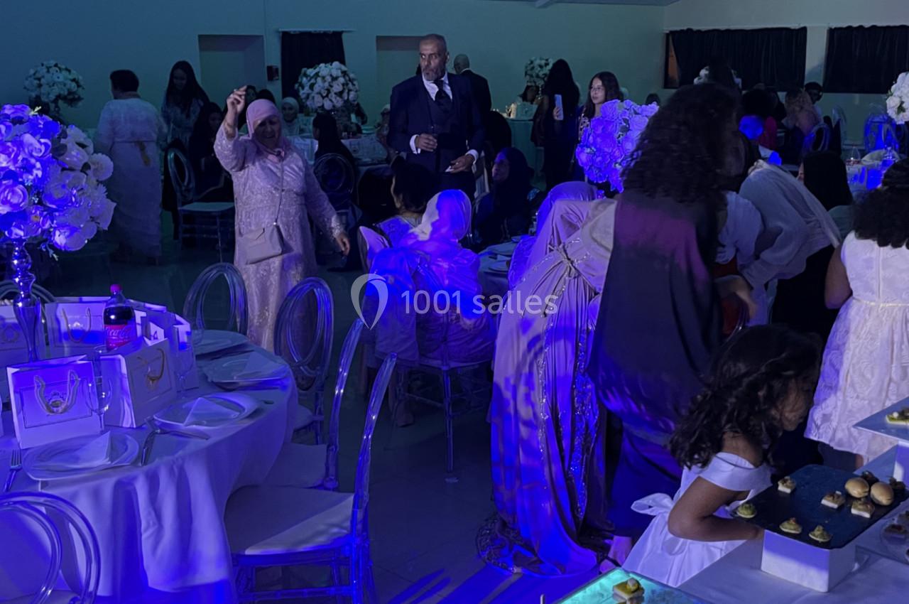 Des invités en tenue élégante se rassemblent dans une salle décorée de fleurs blanches et éclairée par une lumière bleue.