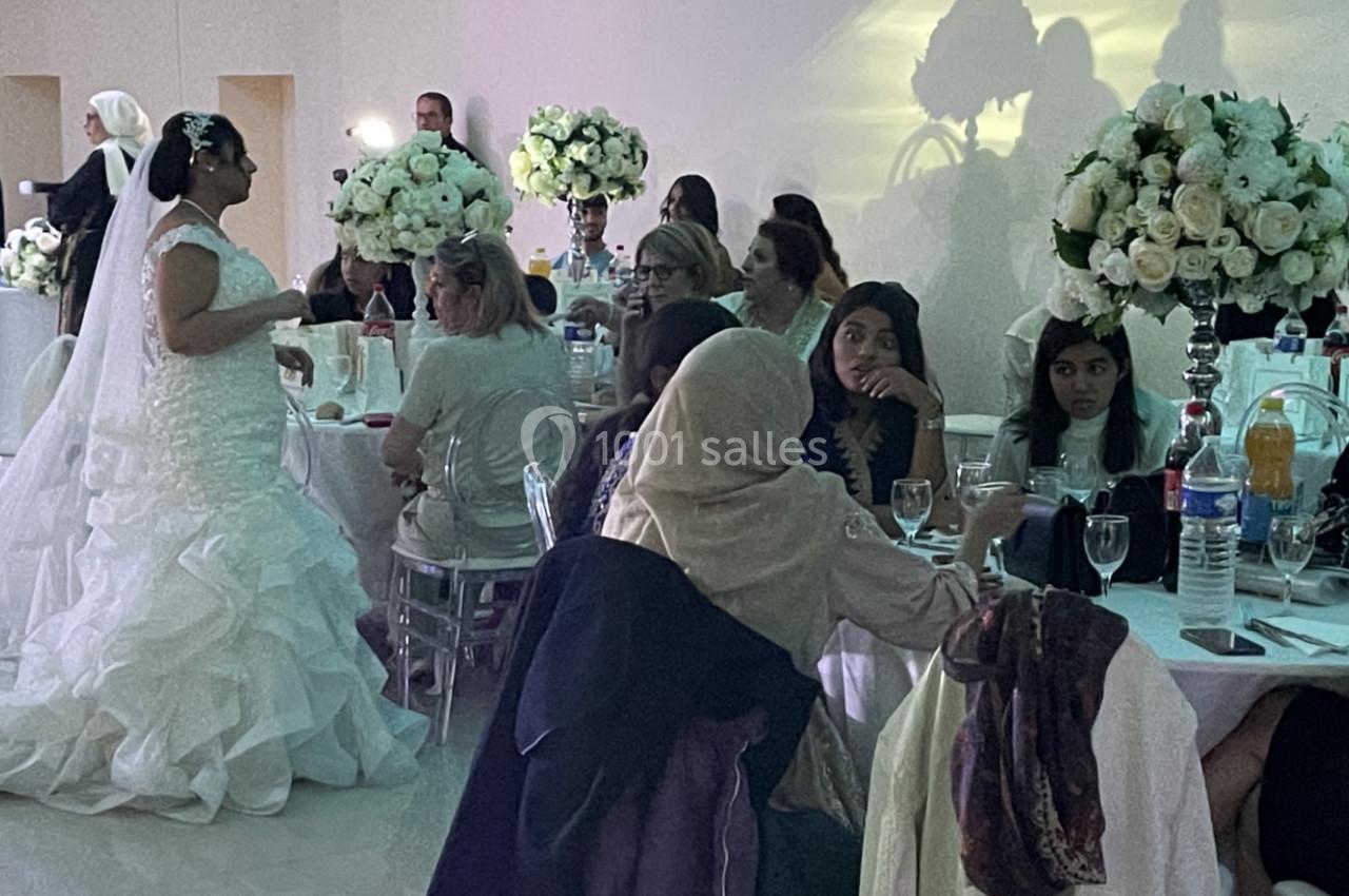 Une mariée en robe blanche échange avec des invités assis à des tables décorées de bouquets de fleurs blanches.