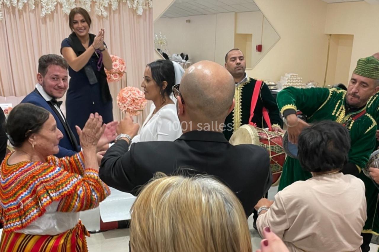Personnes célébrant un mariage dans une salle décorée, avec musiciens en tenue traditionnelle et invités applaudissant.